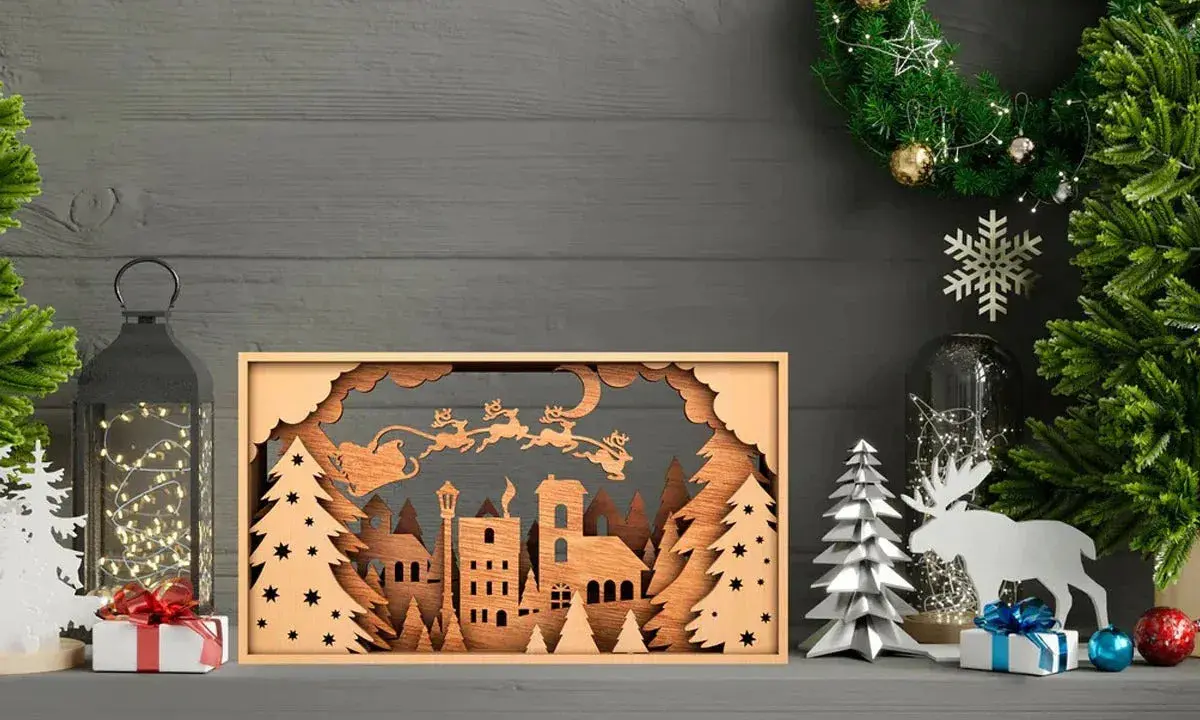 Scena natalizia con Babbo Natale e renne in volo, creata con disegni taglio laser su legno. Un villaggio innevato e alberi completano il quadro.