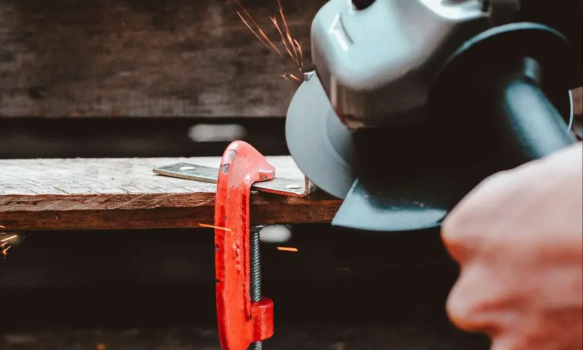 Mano che usa una smerigliatrice angolare per tagliare il ferro, con scintille che volano. Un morsetto rosso tiene fermo il metallo.