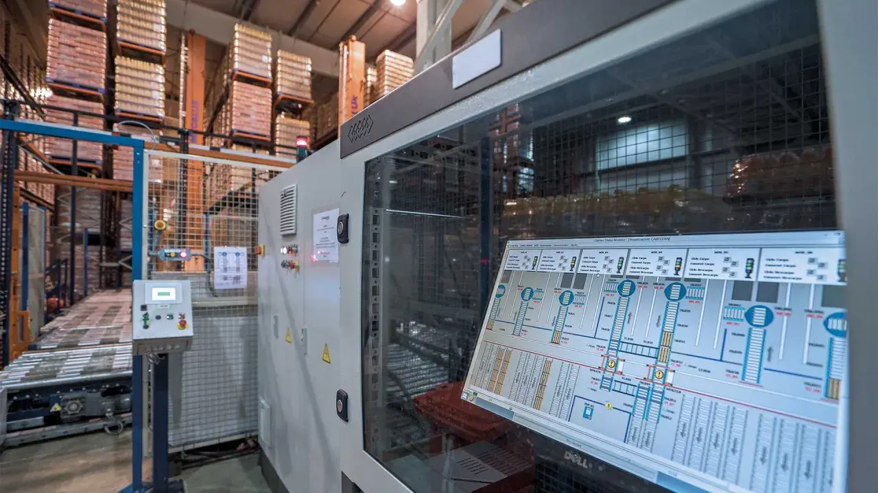 Schermata di un sistema di controllo plc in un magazzino automatizzato, con scaffali carichi di merce sullo sfondo.