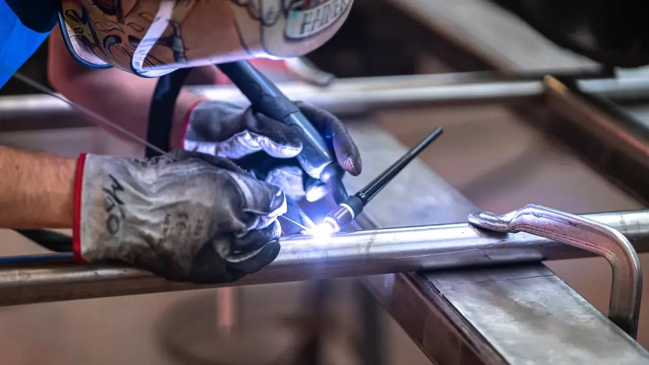 Saldatore con maschera decorata usa torcia TIG per unire tubi metallici, mostrando la differenza tra saldatura a filo e ad elettrodo.