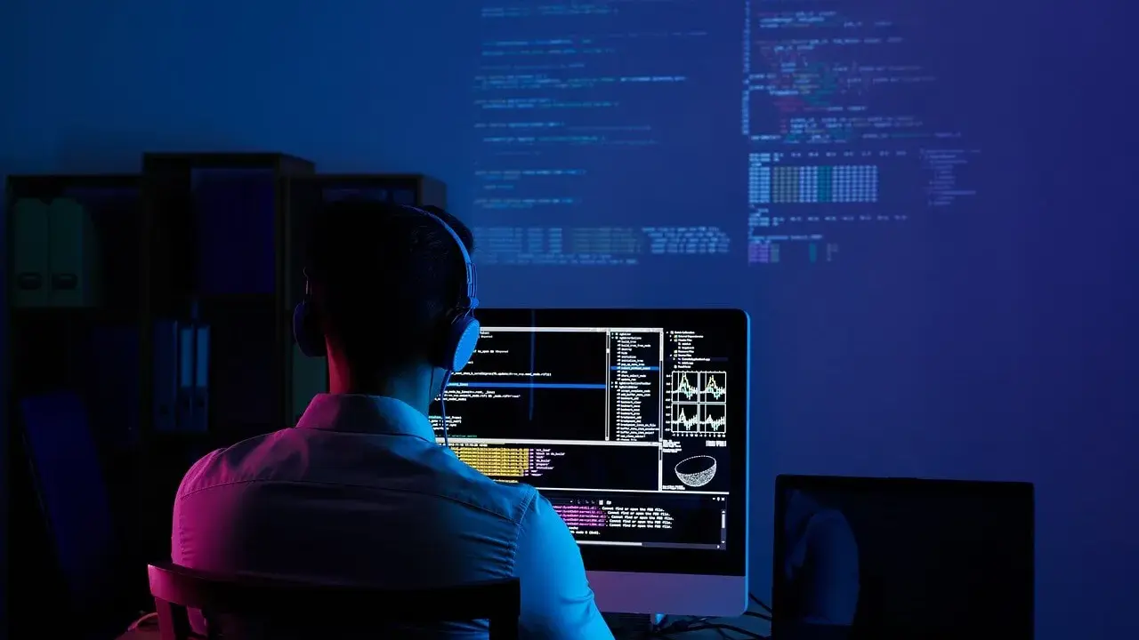 Programmatore con cuffie lavora al suo computer, analizzando dati e codice su più schermi. Il cam software illumina la stanza con luci blu e rosa.