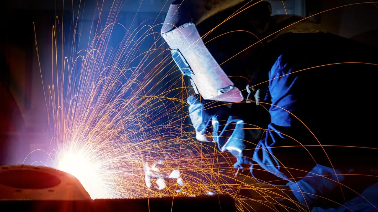 Un saldatore con maschera protettiva esegue lavori di saldatura, creando una pioggia di scintille luminose in un'officina buia.