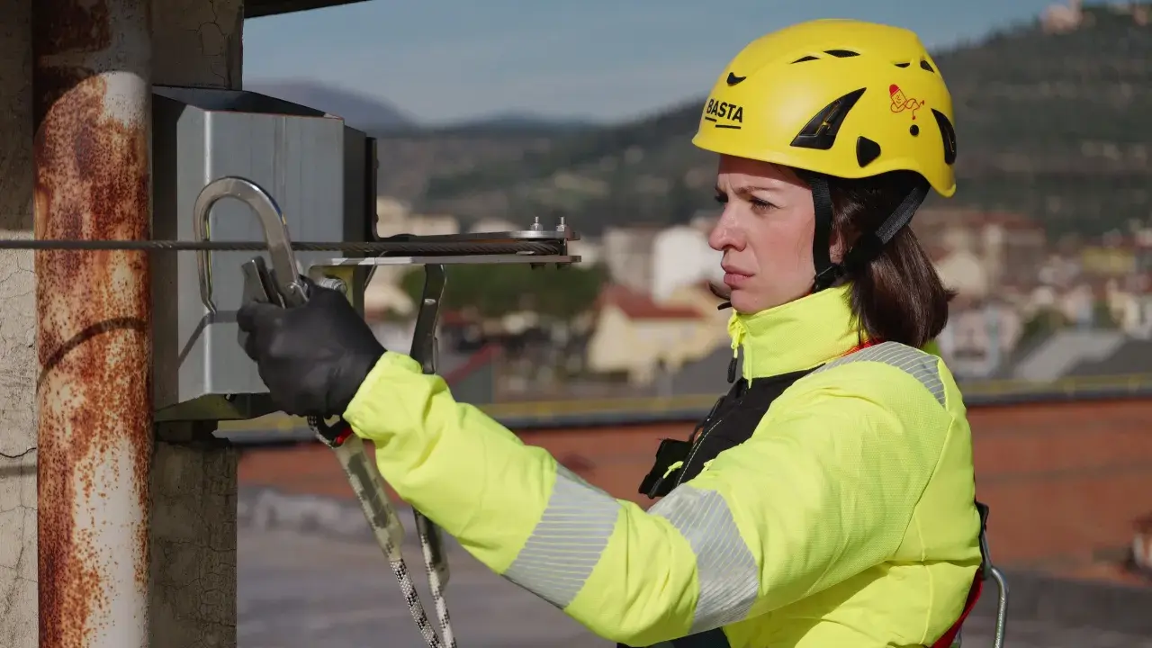 Donna con casco giallo e giacca ad alta visibilità, aggancia un moschettone a un cavo. La sua attrezzatura garantisce la protezione personale in quota.