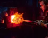 Fabbro con guanti estrae metallo incandescente da forno per la normalizzazione acciaio. Fiamme e calore intenso.