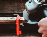 Mano che usa una smerigliatrice angolare per tagliare il ferro, con scintille che volano. Un morsetto rosso tiene fermo il metallo.