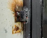 Cerniera arrugginita fissata con cianfrino saldatura su un telaio nero. La vernice bianca circostante è scrostata e macchiata.