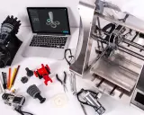 Un programmatore CNC progetta e stampa componenti complessi. Laptop con modello 3D, stampante 3D, parti meccaniche e attrezzi.