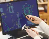 Un programmatore CAD CAM analizza un disegno tecnico su schermo, usando uno stilo e un calibro per misurare dettagli.