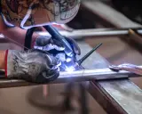 Saldatore con maschera decorata usa torcia TIG per unire tubi metallici, mostrando la differenza tra saldatura a filo e ad elettrodo.