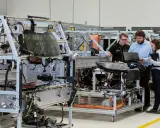 Operai di produzione analizzano schemi su un laptop, lavorando su un telaio di assemblaggio di un veicolo.