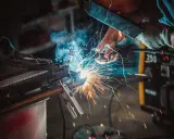 Un operaio con maschera e guanti esegue una saldatura, creando scintille luminose. Il significato della saldatura è unire metalli.