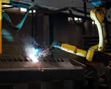 Braccio robotico giallo esegue saldatura, creando scintille luminose su un pezzo metallico grezzo.