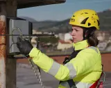 Donna con casco giallo e giacca ad alta visibilità, aggancia un moschettone a un cavo. La sua attrezzatura garantisce la protezione personale in quota.