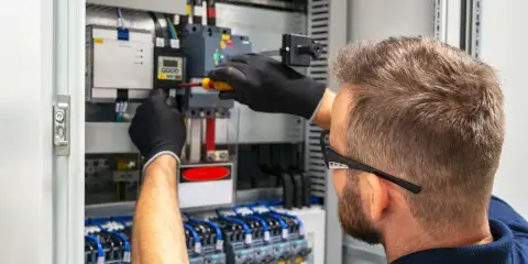 Tecnico con guanti neri lavora su un quadro elettrico, con un cacciavite su un componente. Il suo lavoro con il plc è fondamentale per l'automazione.