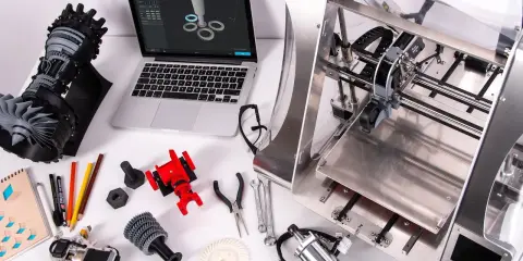 Il disegnatore CAD lavora su un laptop con un modello 3D, circondato da componenti stampati in 3D e strumenti.
