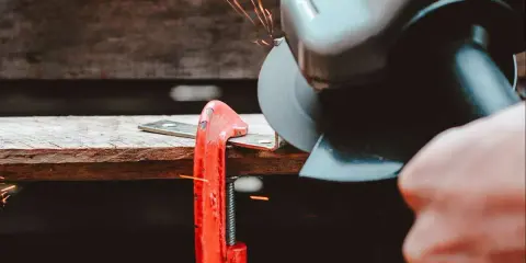 Mano che usa una smerigliatrice angolare per tagliare il ferro, con scintille che volano. Un morsetto rosso tiene fermo il metallo.