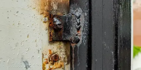 Cerniera arrugginita fissata con cianfrino saldatura su un telaio nero. La vernice bianca circostante è scrostata e macchiata.