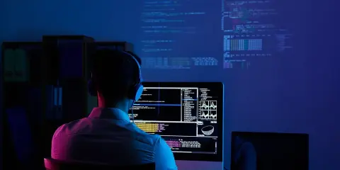 Programmatore con cuffie lavora al suo computer, analizzando dati e codice su più schermi. Il cam software illumina la stanza con luci blu e rosa.