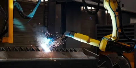 Braccio robotico giallo esegue saldatura, creando scintille luminose su un pezzo metallico grezzo.