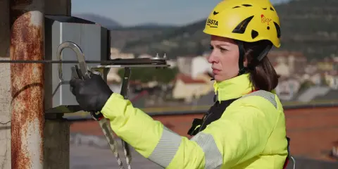 Donna con casco giallo e giacca ad alta visibilità, aggancia un moschettone a un cavo. La sua attrezzatura garantisce la protezione personale in quota.