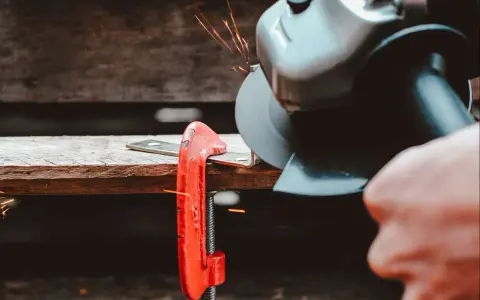 Mano che usa una smerigliatrice angolare per tagliare il ferro, con scintille che volano. Un morsetto rosso tiene fermo il metallo.