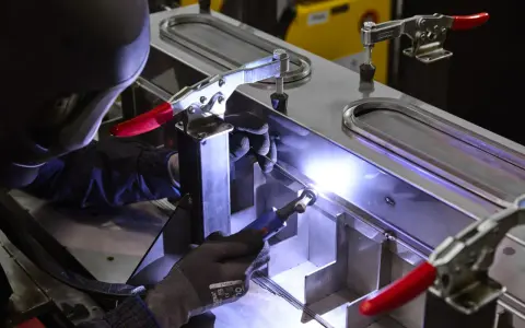 Saldatore esperto esegue saldatura di alluminio su una struttura metallica complessa, illuminata da un bagliore intenso.
