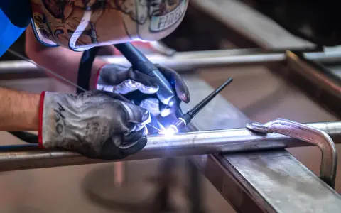 Saldatore con maschera decorata usa torcia TIG per unire tubi metallici, mostrando la differenza tra saldatura a filo e ad elettrodo.