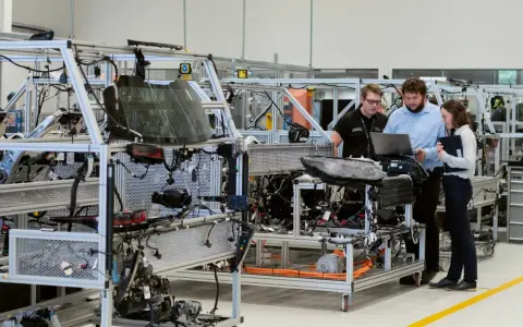 Operai di produzione analizzano schemi su un laptop, lavorando su un telaio di assemblaggio di un veicolo.