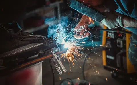 Un operaio con maschera e guanti esegue una saldatura, creando scintille luminose. Il significato della saldatura è unire metalli.