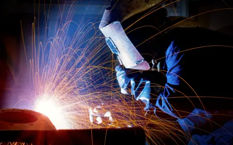 Un saldatore con maschera protettiva esegue lavori di saldatura, creando una pioggia di scintille luminose in un'officina buia.