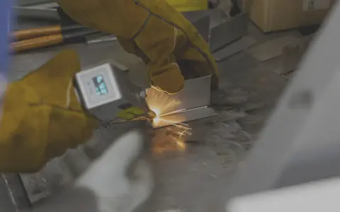 Mani guantate eseguono saldatura laser manuale su metallo, creando scintille luminose.
