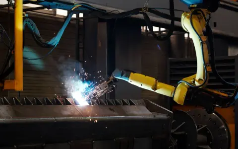 Braccio robotico giallo esegue saldatura, creando scintille luminose su un pezzo metallico grezzo.