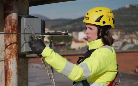 Donna con casco giallo e giacca ad alta visibilità, aggancia un moschettone a un cavo. La sua attrezzatura garantisce la protezione personale in quota.