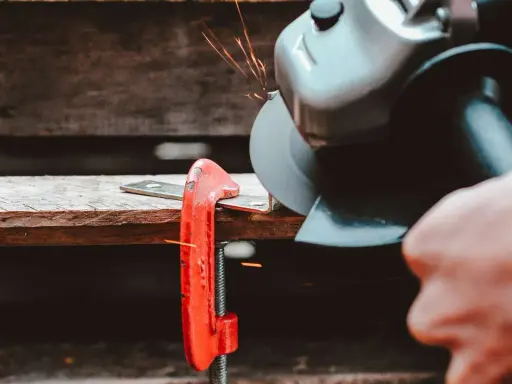 Mano che usa una smerigliatrice angolare per tagliare il ferro, con scintille che volano. Un morsetto rosso tiene fermo il metallo.