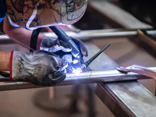 Saldatore con maschera decorata usa torcia TIG per unire tubi metallici, mostrando la differenza tra saldatura a filo e ad elettrodo.