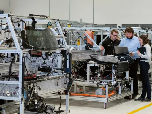 Operai di produzione analizzano schemi su un laptop, lavorando su un telaio di assemblaggio di un veicolo.