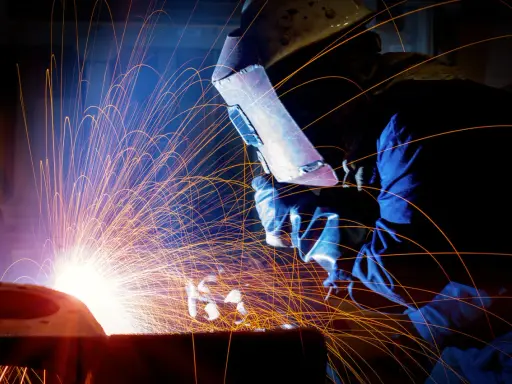 Un saldatore con maschera protettiva esegue lavori di saldatura, creando una pioggia di scintille luminose in un'officina buia.