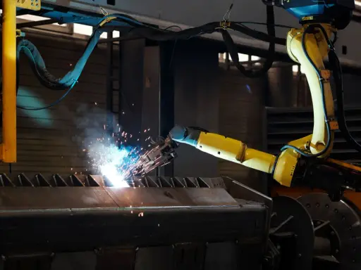 Braccio robotico giallo esegue saldatura, creando scintille luminose su un pezzo metallico grezzo.