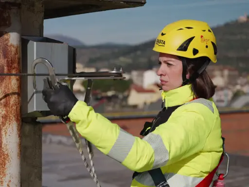 Donna con casco giallo e giacca ad alta visibilità, aggancia un moschettone a un cavo. La sua attrezzatura garantisce la protezione personale in quota.