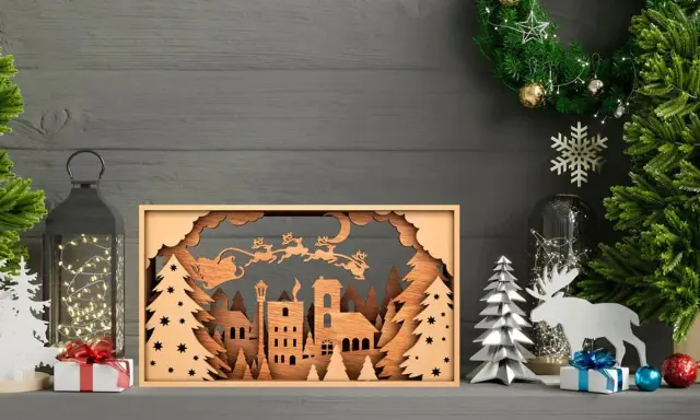 Scena natalizia con Babbo Natale e renne in volo, creata con disegni taglio laser su legno. Un villaggio innevato e alberi completano il quadro.