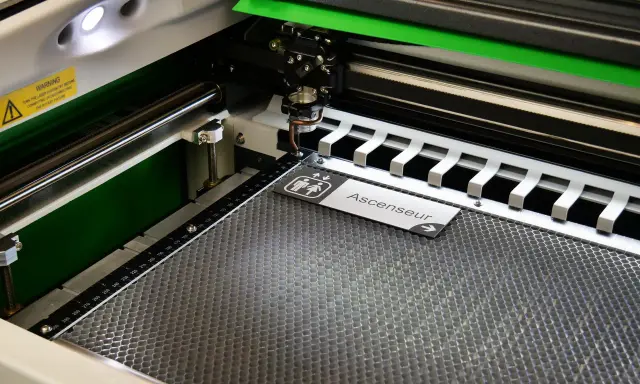 Macchina per taglio laser in azione, che realizza un cartello "Ascenseur" con simboli per bagni. Perfetta per progetti di taglio laser personalizzati.