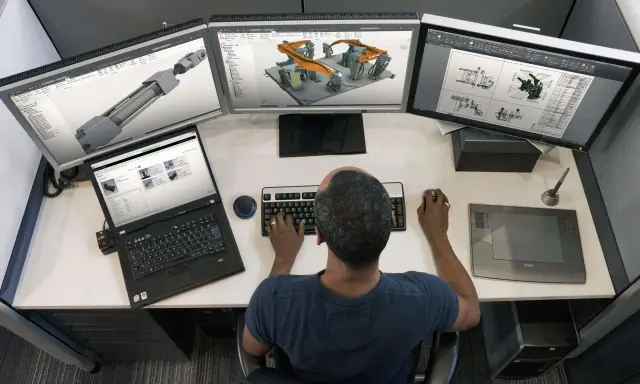 Un disegnatore CAD freelance lavora su un sistema a triplo monitor, creando modelli 3D e disegni tecnici.