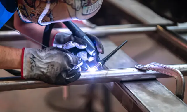 Saldatore con maschera decorata usa torcia TIG per unire tubi metallici, mostrando la differenza tra saldatura a filo e ad elettrodo.