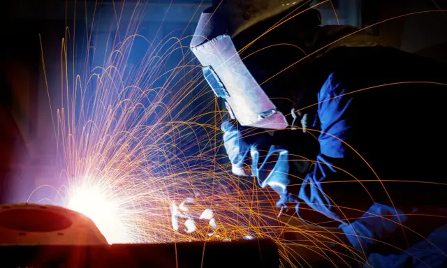 Un saldatore con maschera protettiva esegue lavori di saldatura, creando una pioggia di scintille luminose in un'officina buia.