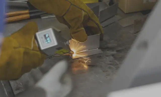 Mani guantate eseguono saldatura laser manuale su metallo, creando scintille luminose.
