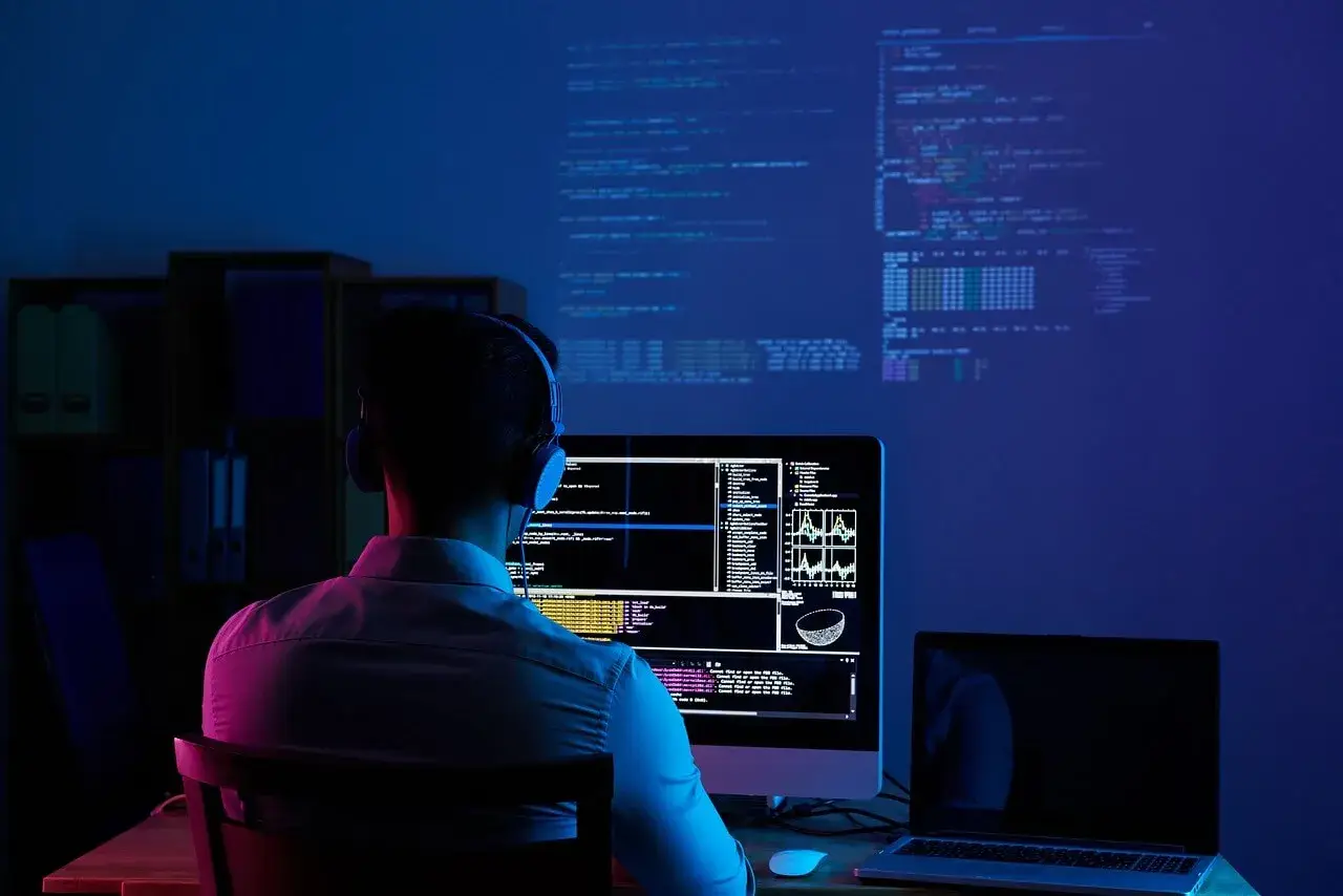 Programmatore con cuffie lavora al suo computer, analizzando dati e codice su più schermi. Il cam software illumina la stanza con luci blu e rosa.