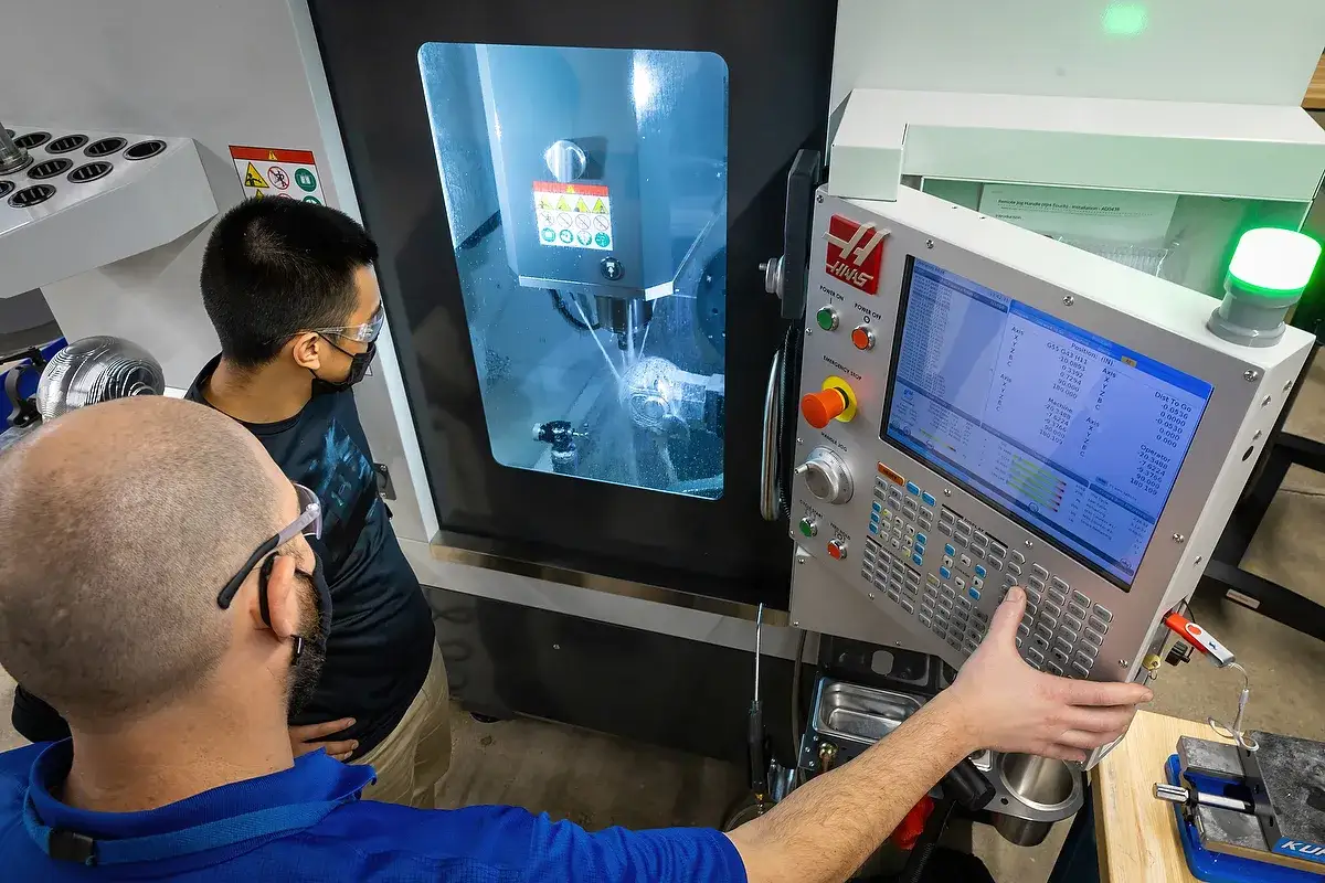 Un programmatore CNC osserva la macchina in azione, mentre un collega controlla i parametri sul pannello di controllo.