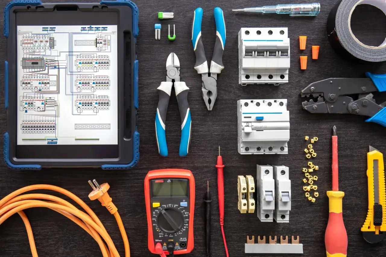 Strumenti elettrici e schema su tablet, tutto rohs compliant. Pinze, cacciaviti, interruttori, multimetro e cavo arancione pronti per un lavoro sicuro.