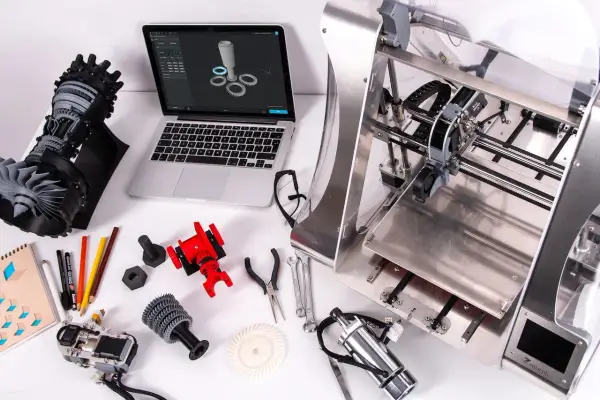 Un programmatore CNC progetta e stampa componenti complessi. Laptop con modello 3D, stampante 3D, parti meccaniche e attrezzi.
