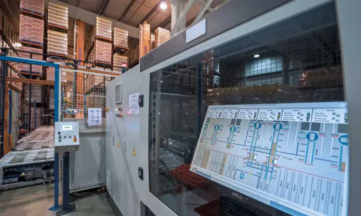 Schermata di un sistema di controllo plc in un magazzino automatizzato, con scaffali carichi di merce sullo sfondo.